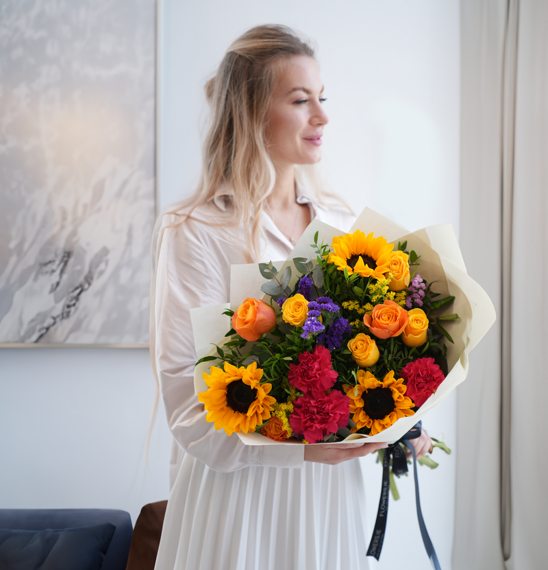 Woman in white dress holding vibrant hand-tied bouquet with sunflowers, orange roses, pink carnations and purple statice