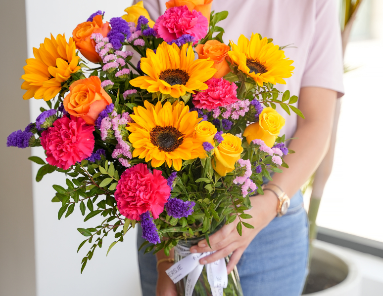 Vibrant hand-tied bouquet with sunflowers, orange roses, pink carnations and purple statice held by woman in casual outfit
