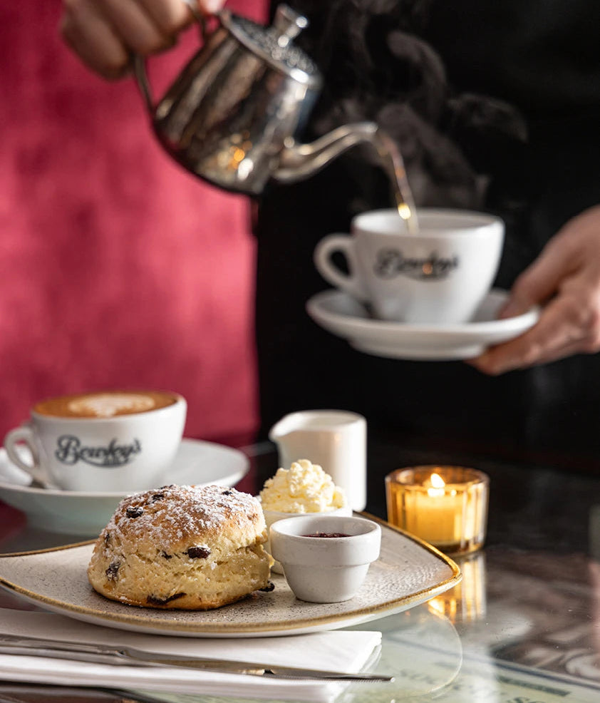 A close-up lifestyle shot of a cafe table setting with a person pouring hot water from a silver teapot into a steaming cup from the brand 'Beaulieu'. In the foreground, a baked scone with powdered sugar and a small bowl of jam sit on a plate on a glass table.