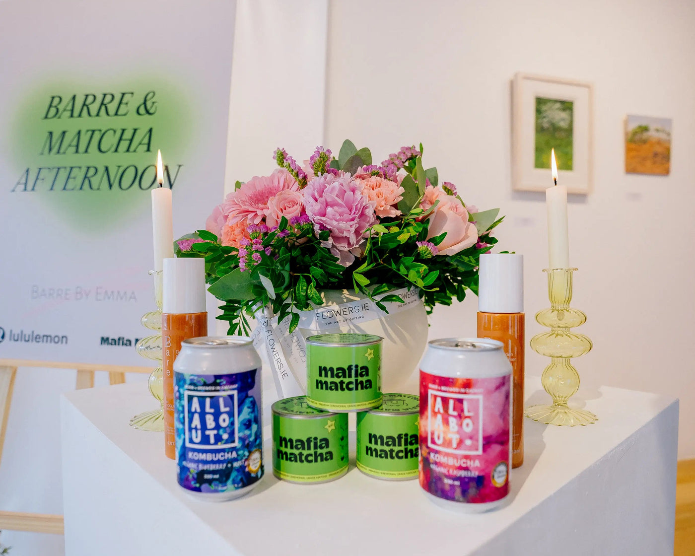 A white display table in a gallery setting showcasing various scented products from Mafia Matcha, including cans and bottles, alongside a vase of pink flowers and a candle. A large sign in the background reads 'lululemon BARRE & MATCHA AFTERNOON'