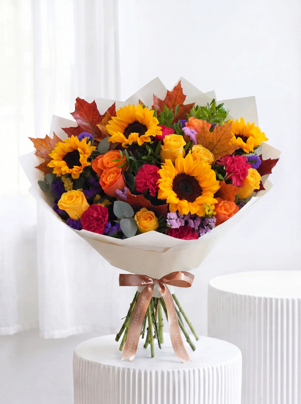 A vibrant autumn floral bouquet featuring sunflowers, orange roses, pink carnations, purple filler flowers, and colorful fall leaves, wrapped in white paper with a rose-gold ribbon and tied to a white pedestal.