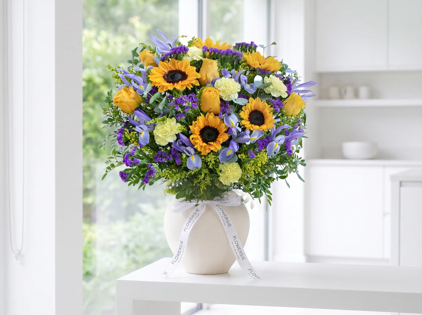 A vibrant sunflower and purple flower bouquet in a white ceramic vase, styled on a white table in a bright, modern kitchen setting